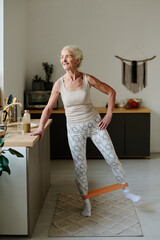 Happy senior woman in activewear stretching elastic band by her legs while holding by kitchen...