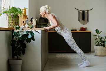 Side view of aged retired woman in activewear doing press ups against kitchen counter during...