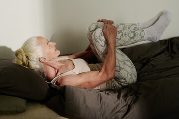 Senior woman in leggins and tanktop lying on bed and holding legs bent in knee against chest while working out after sleep