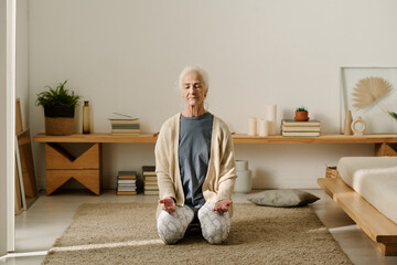 Senior woman in casualwear sitting on her knees on the floor of bedroom and keeping eyes closed while practicing yoga exercise - obrazy, fototapety, plakaty