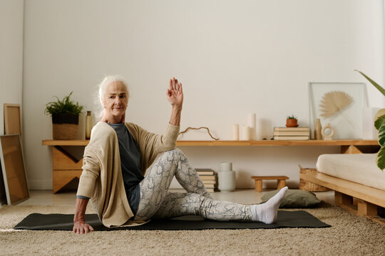Aged active woman in casualwear sitting on the floor of bedroom and practicing yoga exercises while keeping one hand raised