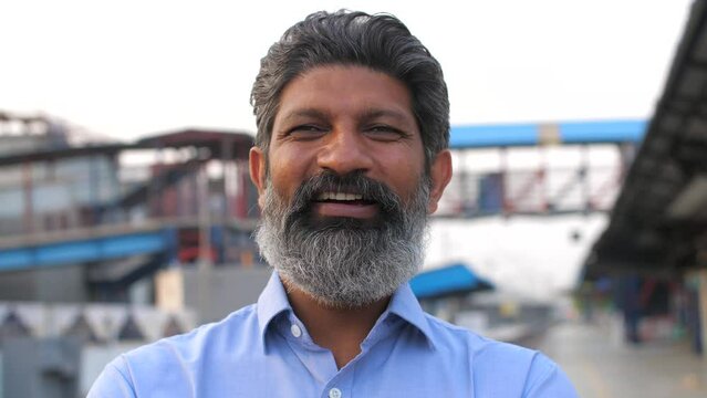 A closeup headshot of a middle-aged Indian businessman standing outdoors at a railway platform - headshot portrait. A confident Indian man with salt and pepper look smiling and posing for the camer...