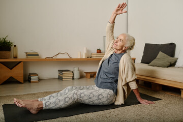 Retired woman in activewear sitting on mat on the floor of bedroom and doing side bends with raised...