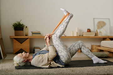 Side view of elderly active woman practicing exercises with resistance band while lying on mat in...