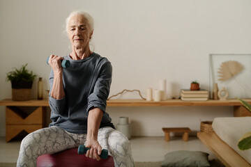 Aged woman in activewear sitting on fitball and practicing physical exercise for arm muscles with...