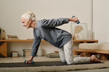 Side view of senior active woman in leggins and pullover standing on knees on the floor of bedroom and doing exercise with dumbbells - obrazy, fototapety, plakaty