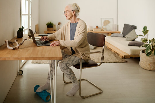 Senior woman in casualwear and eyeglasses using massage roller under table while sitting on chair in front of laptop screen