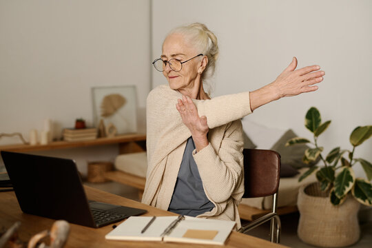 Senior woman in eyeglasses looking at screen of laptop and doing exercise for stretching while sitting by table and working with online data