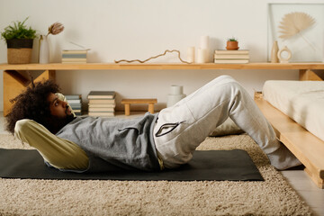 Active young man in sweatpants and hoodie doing sit ups while lying on black mat during morning workout on the floor of bedroom - obrazy, fototapety, plakaty