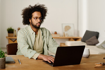 Young serious multiethnic male freelancer or student looking at laptop screen and typing while...