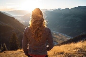  the back of a woman's head as she stands at the top of a mountain overlooking a valley with mountains in the background and the sun shining on the horizon. - obrazy, fototapety, plakaty