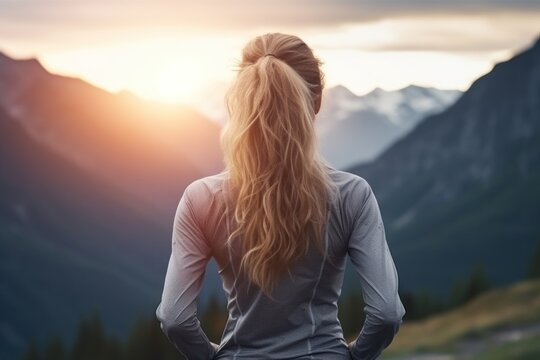  the back of a woman's head as she stands in front of a mountain range with the sun behind her and her back to the camera, with mountains in the background.
