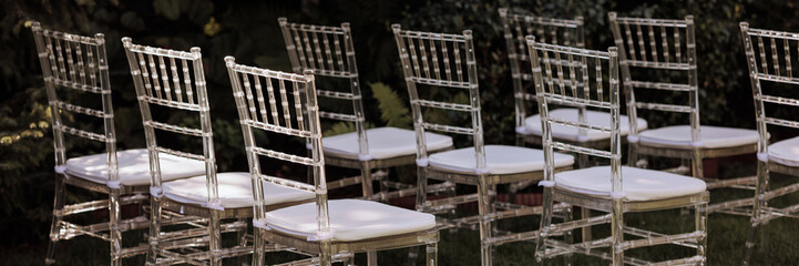 Row of elegant, transparent chairs with white cushions set outdoors in soft sunlight. Concept: outdoor events, modern design, or garden parties. Banner with copy space.