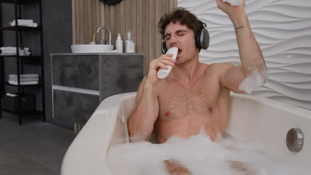 Carefree positive silly funny Caucasian naked man male guy alone relaxing in bathroom in warm bath water enjoy home resting singing song in shower gel like microphone listening to music in headphones