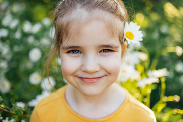 Child portrait of cute little blue-eyed girl with flower of chamomile behind her ear in park or...