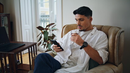 Serious guy enjoying whiskey cola home interior. Relaxed man reading smartphone