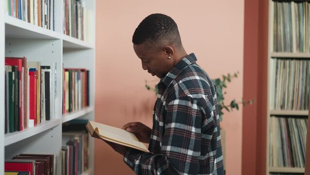 Man reads book standing next to shelf. Thoughtful dark-skinned bookworm dips into literature standing in premise of college library. Reading as hobby