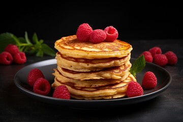  a stack of pancakes on a black plate with raspberries on the side and a leafy branch of mint on the other side of the stack of pancakes.