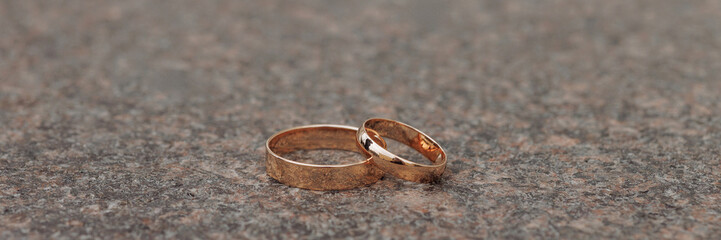 Two golden wedding bands, simple and without gemstones, are placed closely together on a textured stone or concrete, surface. Concept for wedding, love, and commitment. Banner with copy space.