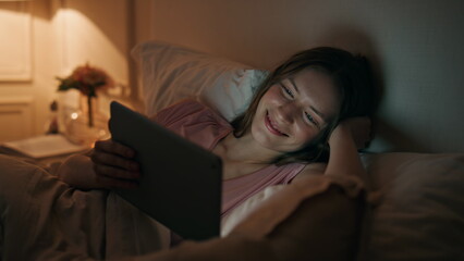 Girl watching tablet online at night closeup. Smiling woman resting bed enjoying