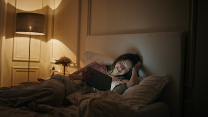 Relaxed girl looking tablet in cozy bed at night. Smiling happy woman watching