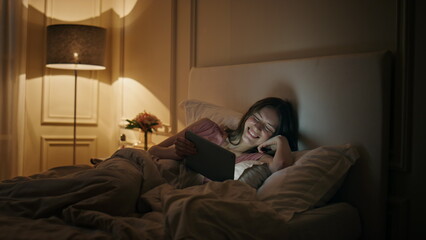Cheerful woman watching tablet in bed. Happy female laughing movie in evening.