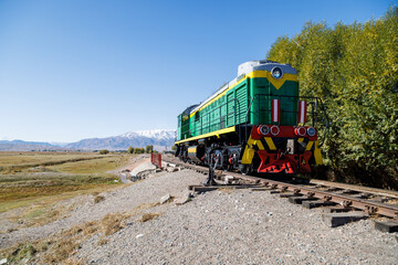 green diesel locomotive crossing small bridge with mountains in the background at summer day in...