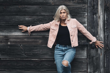 Older woman in her fifties in front of an old wooden wall, dressed in a pink denim jacket, blue...