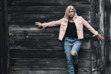 Older woman in her fifties in front of an old wooden wall, dressed in a pink denim jacket, blue jeans and a black T-shirt, in a retro style