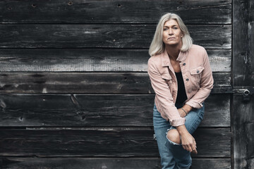 Older woman in her fifties in front of an old wooden wall, dressed in a pink denim jacket, blue jeans and a black T-shirt, in a retro style