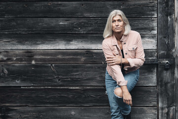 Older woman in her fifties in front of an old wooden wall, dressed in a pink denim jacket, blue jeans and a black T-shirt, in a retro style