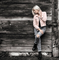Older woman in her fifties in front of an old wooden wall, dressed in a pink denim jacket, blue...