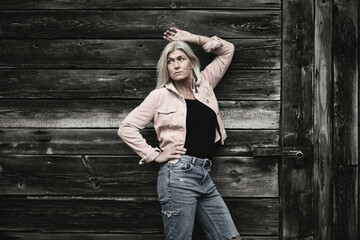 Older woman in her fifties in front of an old wooden wall, dressed in a pink denim jacket, blue jeans and a black T-shirt, in a retro style