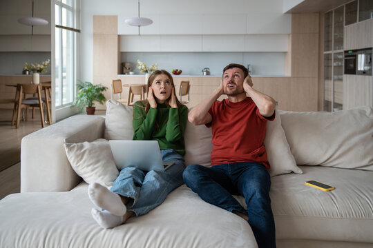 Annoyed unhappy couple sitting on sofa with vexed face expression, looking upward, closing ears because of loud sound, noise from neighboring flat. Emotional stress, discomfort caused by neighborhood