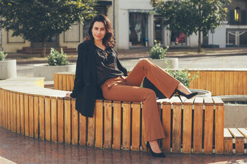 A relaxed yet stylish woman sits comfortably on a curved wooden bench in an urban plaza, sporting a...