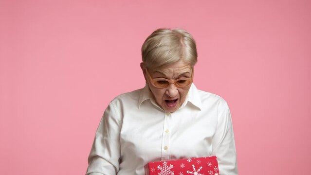 Pleasantly surprised senior lady in stylish spectacles white shirt after opening box wrapped in red paper with snowflakes, standing over pink background. High quality 4k footage