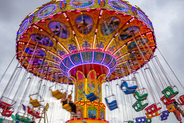 Detalhe de um brinquedo gigante de rotação no parque de diversões com poucas pessoas, rodando em baixa velocidade, em um dia nublado. Foto feita de baixo para cima.