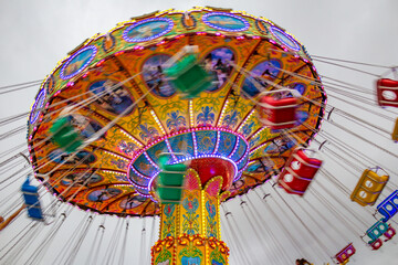 Um brinquedo de rotação, grande e colorido, no parque de diversões com poucas pessoas, em um dia nublado. Foto feita de baixo para cima. - obrazy, fototapety, plakaty
