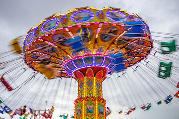 Um brinquedo gigante de rotação, com várias cadeiras no alto, borradas pela velocidade do movimento, no parque de diversões com poucas pessoas, em um dia nublado. Foto feita de baixo para cima. - obrazy, fototapety, plakaty