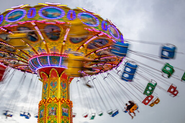 Um brinquedo gigante de rotação, com várias cadeiras no alto, borradas pela velocidade do movimento, no parque de diversões com poucas pessoas, em um dia nublado. Foto feita de baixo para cima. - obrazy, fototapety, plakaty
