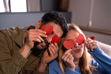 Smiling couple holding heart shapes over their eyes while sitting on the couch at home .Valentine's...
