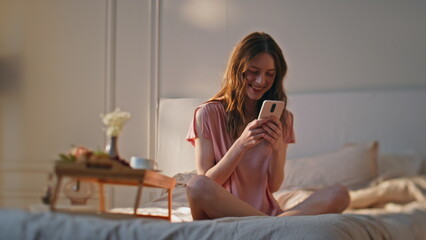 Cozy girl typing mobile phone in hotel bed. Dreamy happy woman get good news