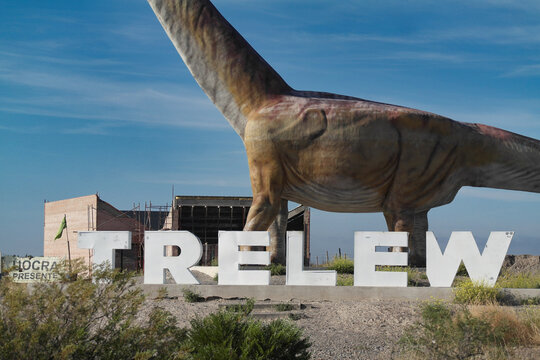 The image shows a billboard that advertises the presence of the world&rsquo;s largest dinosaur in Trelew. The main focus is a large white billboard with black and blue text, set against a clear sky