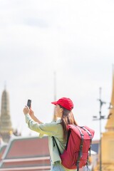 Young asian woman traveler with phone capturing moments and exploring Wat Phra Kaew and The Grand Palace in a happy and casual style during a sunny day - obrazy, fototapety, plakaty