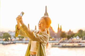 Young Asian Woman Traveler Taking a Selfie While Enjoying The Sunset Moments of Wat Arun by the Chao Phraya Riverbank in Bangkok, Thailand - obrazy, fototapety, plakaty