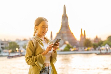 Naklejka premium Young Asian Woman Traveler Holding Phone While Enjoying The Sunset Moments of Wat Arun by the Chao Phraya Riverbank in Bangkok, Thailand