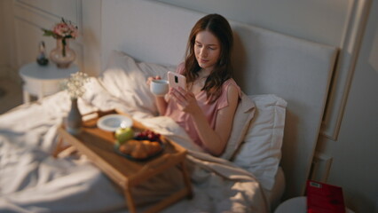 Obraz na płótnie Canvas Calm girl enjoying breakfast drinking coffee in bed. Relaxed female surfing web