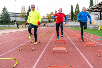 Obraz premium Three unrecognizable senior runners on a track doing high knee drills over yellow and red hurdles for agility