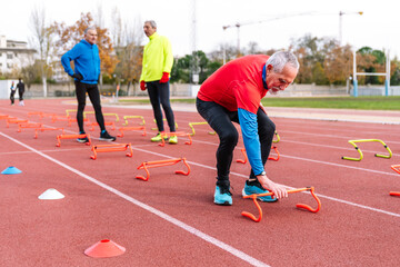 Fototapeta premium Senior man in red bends over to adjust training cone, with two others discussing the exercise session