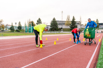 Elderly sportsmen on an athletic track, one with a trolley of cones, another bending to place cones.